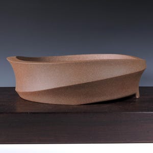 May include: A brown ceramic bonsai pot with a unique, curved design. The pot has a textured surface and a wide, open top. The sides curve inward, creating an elegant, modern look. The pot is designed for planting small trees or plants.