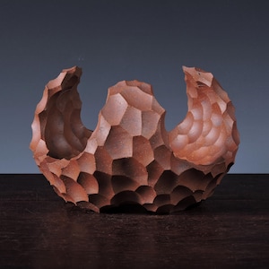 May include: A reddish-brown ceramic bowl with a unique, textured surface. The bowl has a geometric pattern of raised shapes and two curved openings on opposite sides. The overall shape is reminiscent of a stylized landscape or a natural form.