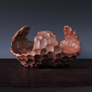 May include: A handcrafted, reddish-brown wooden bowl with a unique, textured surface. The bowl's exterior features a geometric, honeycomb-like pattern, and the rim has an organic, undulating shape. The bowl is displayed on a dark wooden surface.