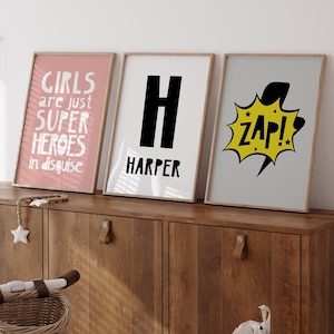 May include: Three framed prints with a pink, white, and gray background. The pink print says "Girls are just super heroes in disguise". The white print has a black "H" and the word "Harper" below it. The gray print has a yellow lightning bolt with the word "Zap!" inside.