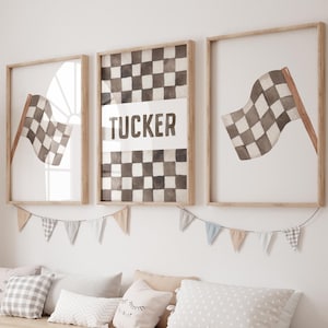 May include: Three framed watercolour prints with a racing theme. Each print features a checkered flag in shades of grey and brown. The central print has the name "TUCKER". A garland of small pennants hangs below the frames.