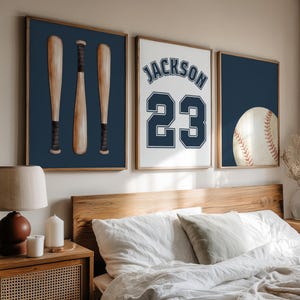 May include: A set of three framed wall art prints featuring baseball themes. The left print shows three wooden bats on a navy blue background. The center print displays the name JACKSON and the number 23 in navy blue. The right print shows a baseball.