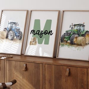 Może przedstawiać: Trzy oprawione wydruki z akwarelowymi ilustracjami traktor&oacute;w i bel siana. Jeden wydruk przedstawia niebieski traktor, drugi zielony traktor, a środkowy wydruk ma imię "mason" z zieloną literą "M". Ramki są jasnobrązowe.