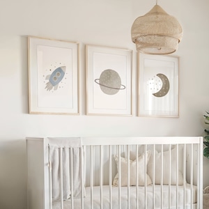 May include: A white crib with a white mattress and a white blanket. There are three framed prints on the wall above the crib, each featuring a different space-themed illustration. A large woven pendant light hangs above the crib. A small potted plant sits on the floor next to the crib.