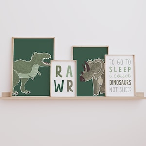 Op de afbeelding: Vier ingelijste prints met een dinosaurus thema. De prints tonen een groene Tyrannosaurus Rex, een groene Triceratops, het woord "RAWR" in groene en bruine letters, en de tekst "TO GO TO SLEEP I COUNT DINOSAURS NOT SHEEP" in groene en bruine letters.