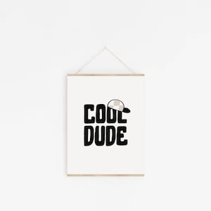 May include: A white framed print with black text that reads "COOL DUDE" with a small beige and white checkered baseball cap above the word "COOL".