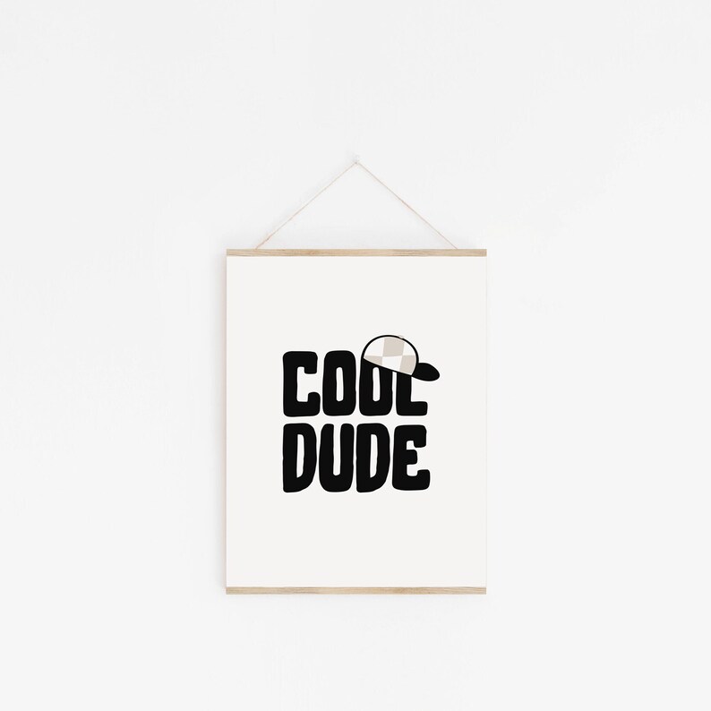 May include: A white framed print with black text that reads "COOL DUDE" with a small beige and white checkered baseball cap above the word "COOL".