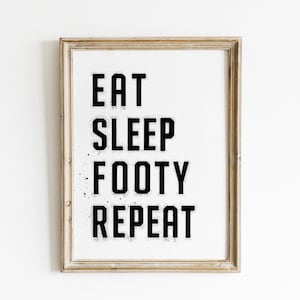 Puede incluir: Impresión enmarcada con las palabras "EAT SLEEP FOOTY REPEAT" en letras negras en negrita sobre un fondo blanco. El marco es de color marrón claro y el texto tiene un efecto desgastado y salpicado.
