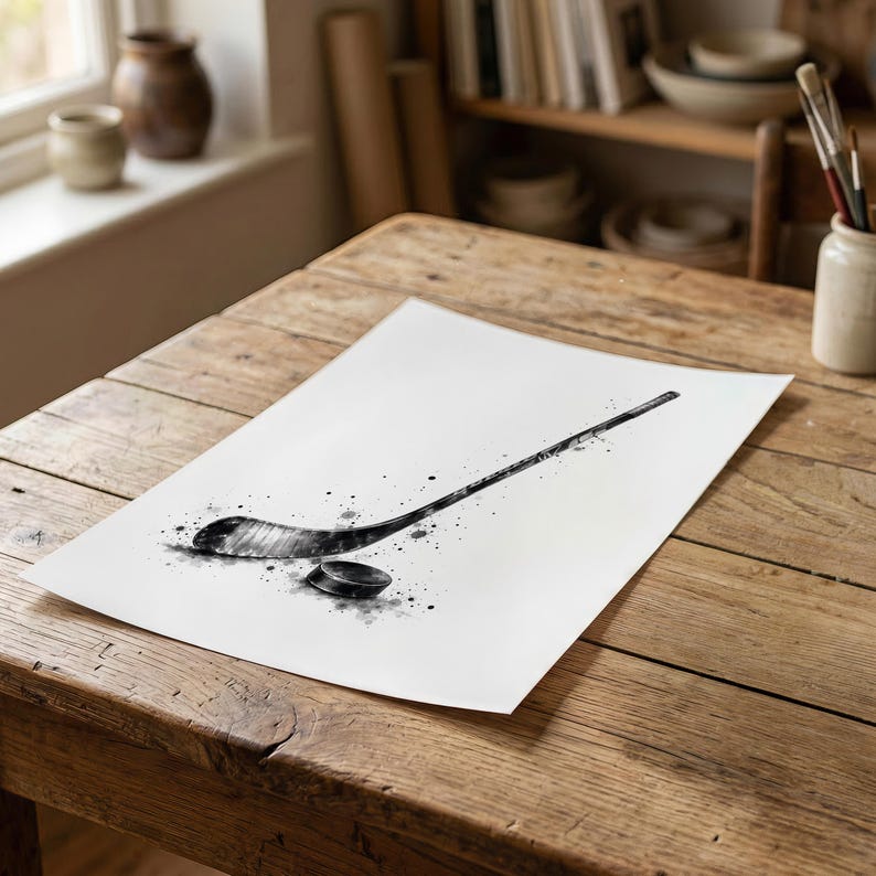 Pode incluir: Uma ilustra&ccedil;&atilde;o em estilo aguarela a preto e branco sobre papel, representando um stick de h&oacute;quei e um disco. A obra apresenta salpicos de tinta expressivos e v&aacute;rios tons de cinzento, criando uma sensa&ccedil;&atilde;o din&acirc;mica de movimento e textura.