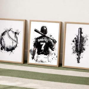 May include: Three framed wall art prints featuring black and white watercolor-style illustrations of baseball equipment. The left print shows a baseball, the center print depicts a player with the name BROOKS and number 7, and the right print shows a bat.