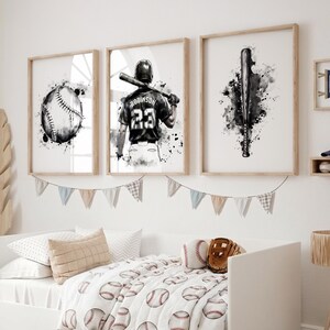 May include: Three framed wall art prints featuring black and white watercolor illustrations of a baseball, a player wearing a jersey with the number 23 and the name Robinson, and a baseball bat, displayed above a bed with baseball-themed bedding.