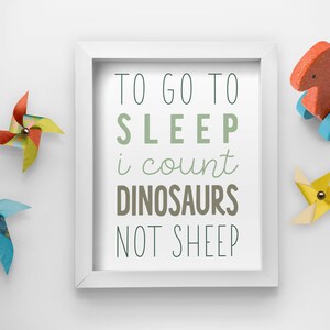 Op de afbeelding: Witte ingelijste print met de tekst "Om te gaan slapen tel ik dinosaurussen, geen schapen". De tekst is in een groen, bruin en zwart lettertype.