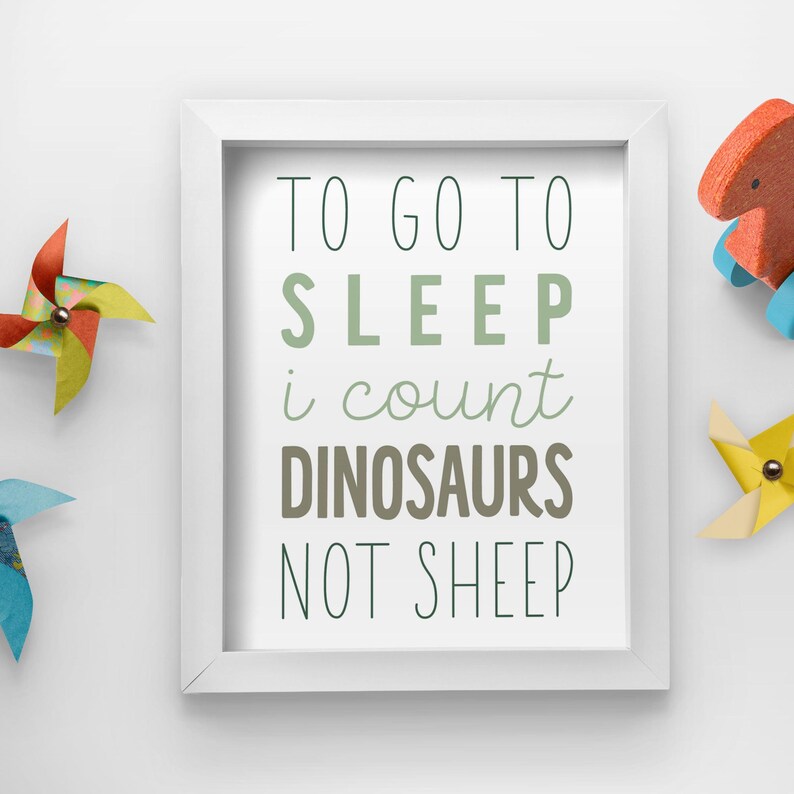 Op de afbeelding: Witte ingelijste print met de tekst "Om te gaan slapen tel ik dinosaurussen, geen schapen". De tekst is in een groen, bruin en zwart lettertype.