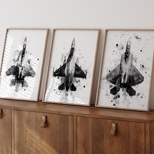 Puede incluir: Tres impresiones enmarcadas en blanco y negro de aviones de combate. Cada impresión presenta un diseño de avión diferente con un fondo salpicado al estilo acuarela. Los marcos son de color madera clara y las impresiones se exhiben en un mueble de madera.
