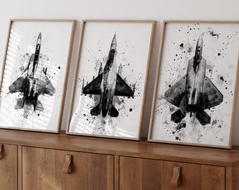 May include: Three framed black and white prints of fighter jets. Each print features a different jet design with a watercolor-style splatter background. The frames are a light wood color, and the prints are displayed on a wooden cabinet.