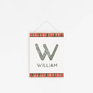 May include: A white sign with a gray watercolor letter W and the name William printed below. The sign is framed with a red and green plaid border.