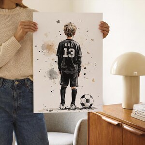 Puede incluir: Una l&aacute;mina art&iacute;stica estilo acuarela que muestra a un ni&ntilde;o visto desde atr&aacute;s, con una camiseta de f&uacute;tbol negra con el nombre SAL y el n&uacute;mero 13 impresos en blanco. El ni&ntilde;o lleva pantalones cortos y calcetines negros junto a un bal&oacute;n.