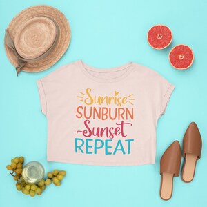 K&ouml;nnte beinhalten: Ein rosa, bauchfreies T-Shirt mit dem Text "Sunrise Sunburn Sunset Repeat" in bunten Buchstaben. Das Shirt liegt auf einem hellblauen Hintergrund mit einem Strohhut, Trauben, einem Glas Wein und zwei Grapefruit-H&auml;lften.