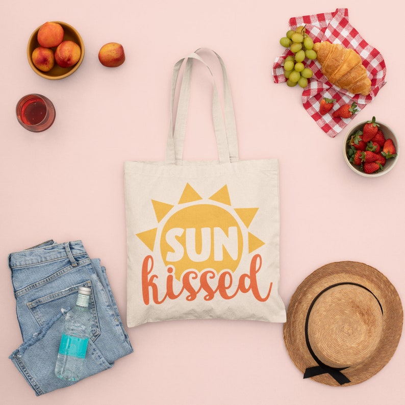K&ouml;nnte beinhalten: Eine wei&szlig;e Canvas-Tragetasche mit dem Text "SUN kissed" in Orange und Gelb. Die Tasche zeigt eine stilisierte Sonnengrafik mit orangefarbenen und gelben Strahlen.