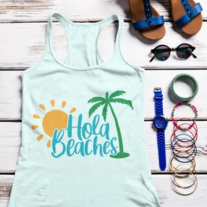 K&ouml;nnte beinhalten: Ein hellblaues Tanktop mit einem Palmen- und Sonnenmotiv und dem Text "Hola Beaches". Das Tanktop liegt flach auf einer wei&szlig;en Holzoberfl&auml;che mit einem Paar blauer Sandalen, einer Sonnenbrille, einer blauen Uhr und mehreren Armb&auml;ndern.