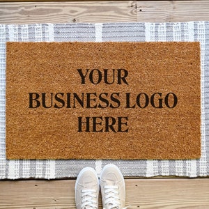 May include: A brown coir doormat with white fringe and a gray and white striped border. The doormat has the text "YOUR BUSINESS LOGO HERE" printed on it.