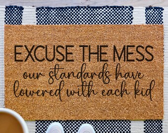 May include: A doormat with the text "EXCUSE THE MESS our standards have lowered with each kid." The mat is made of brown cork with a black and white striped border and fringe. A cup of coffee and a pair of sneakers are visible.