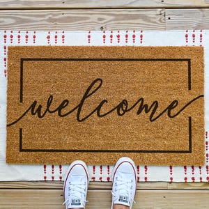 May include: A brown coir doormat with a white border and red accents. The doormat has the word "welcome" written in black cursive script.