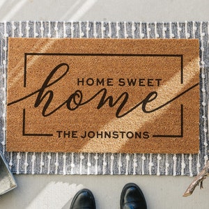 May include: A brown coir doormat with the text "Home Sweet Home" in a black cursive font. The text "The Johnstons" is also printed in black below the main text.