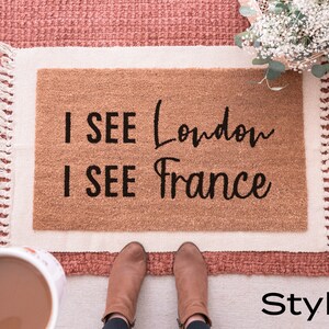May include: A brown doormat with white fringe and the text "I SEE London I SEE France" in black lettering. The doormat is on a pink rug with a bouquet of white flowers in the background.