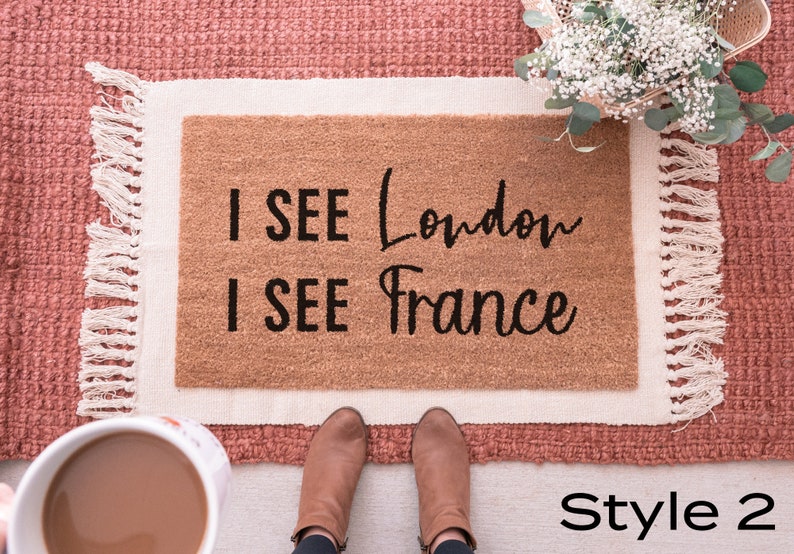 May include: A brown doormat with white fringe and the text "I SEE London I SEE France" in black lettering. The doormat is on a pink rug with a bouquet of white flowers in the background.