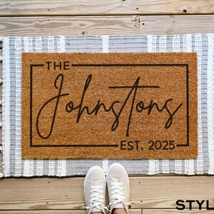 May include: A brown coir doormat with white fringe. The doormat has a black frame with the text "The Johnstons Est. 2025" printed on it.