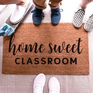 May include: A brown doormat with the text "home sweet classroom" in black lettering.  A group of people stand on the doormat with their shoes on.