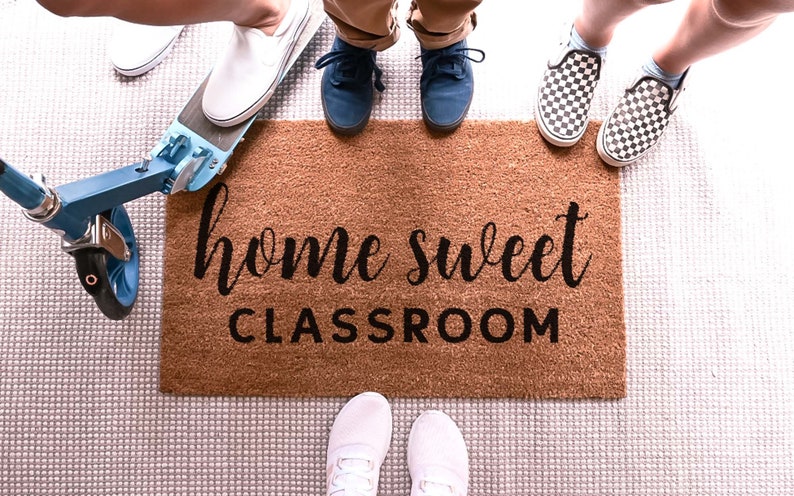 May include: A brown doormat with the text "home sweet classroom" in black lettering.  A group of people stand on the doormat with their shoes on.