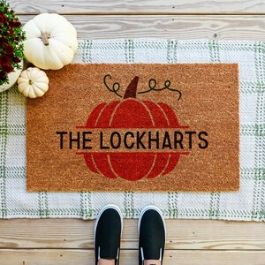 May include: A brown coir doormat with a red pumpkin design and the text "The Lockharts". The doormat has a green and white checkered pattern with fringe.