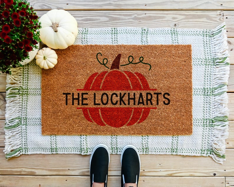 May include: A brown coir doormat with a red pumpkin design and the text "The Lockharts". The doormat has a green and white checkered pattern with fringe.