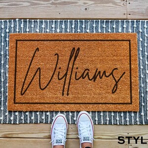 May include: A brown coir doormat with a white border and the name "Williams" written in black cursive script. The doormat is on a blue and white patterned rug.