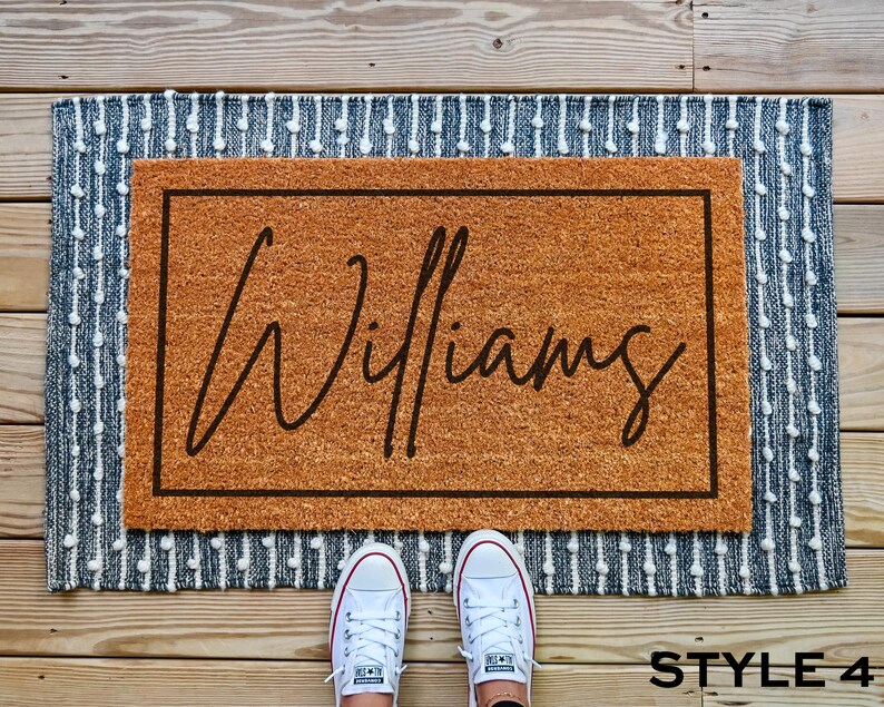 May include: A brown coir doormat with a white border and the name "Williams" written in black cursive script. The doormat is on a blue and white patterned rug.