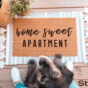 May include: A brown coir doormat with the text "home sweet apartment" in black lettering. The doormat is on a white and gray striped rug with white tassels.