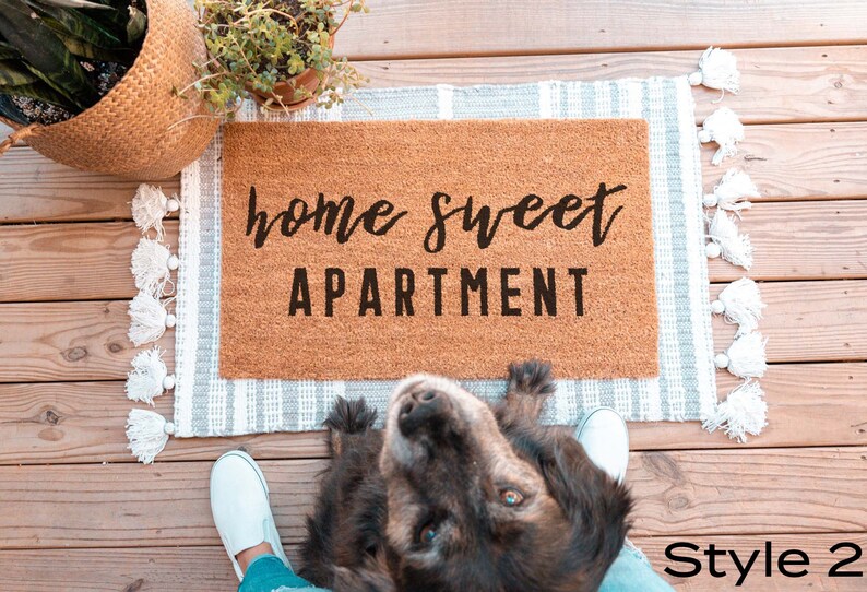 May include: A brown coir doormat with the text "home sweet apartment" in black lettering. The doormat is on a white and gray striped rug with white tassels.
