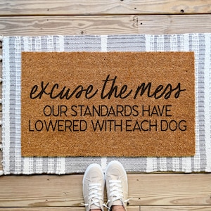 Puede incluir: Una estera de coco marr&oacute;n con el texto "excuse the mess our standards have lowered with each dog" en letras negras. La estera est&aacute; sobre una alfombra rayada blanca y gris con borlas.