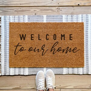 May include: A brown coir doormat with the text "WELCOME to our home" in black lettering. The doormat is surrounded by a white and gray striped rug with tassels.