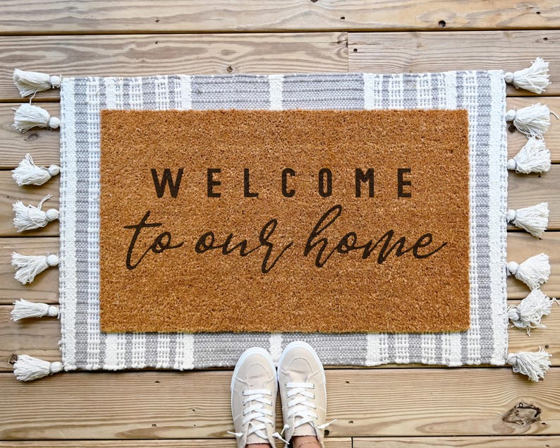 May include: A brown coir doormat with the text "WELCOME to our home" in black lettering. The doormat is surrounded by a white and gray striped rug with tassels.