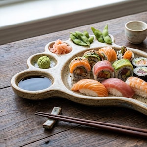 May include: A foot-shaped ceramic sushi platter filled with various sushi rolls, nigiri, and garnishes. Includes wasabi, pickled ginger, soy sauce, and edamame. Chopsticks and a sake set are also present on a wooden table.