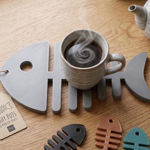 May include: A gray fish-shaped coaster holds a mug of steaming coffee. The coaster is next to a tag that reads "FISHY POTS 3D PRINTED CRAFT". Additional coasters in black, copper, and teal are visible, along with a teapot and a spool of filament.