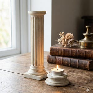 Può includere: Un portacandele a colonna scanalata color crema con base quadrata è accanto a un piccolo portacandele rotondo con una candela da tè accesa. Gli oggetti sono su un tavolo di legno, con libri antichi e un oggetto in ottone sullo sfondo.