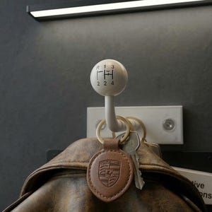 May include: A white gear shift knob-shaped wall hook with black markings for gears. A leather keychain with a Porsche emblem and keys hang from the hook. A brown leather jacket is partially visible below the hook.