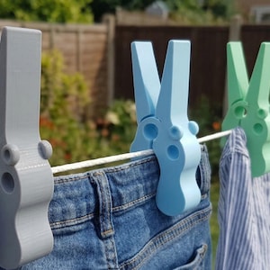 Könnte beinhalten: Eine Wäscheleine mit mehreren großen, dekorativen Wäscheklammern in Grau-, Blau- und Grüntönen. Die Wäscheklammern halten Jeans und Hemden fest. Der Hintergrund ist eine verschwommene Ansicht eines Gartens.