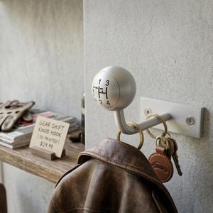 Op de afbeelding: Een zilveren versnellingspookknophaak gemonteerd op een witte metalen plaat, met sleutels en een bruine leren jas die eraan hangt. De versnellingspookknop heeft de nummers 1, 2, 3 en 4 erop gedrukt. De haak is bevestigd aan een betonnen muur.