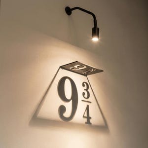 May include: A black metal sign with the numbers "9 3/4" in a stylized font, mounted on a wall. The sign is illuminated by a black, adjustable spotlight. The sign is a reference to a fictional location.