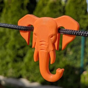 Peut inclure: Un crochet en forme de tête d'éléphant orange vif imprimé en 3D avec une surface texturée. Les oreilles de l'éléphant sont placées sur un cordon tressé noir, permettant à la trompe de pendre vers le bas comme un crochet fonctionnel.